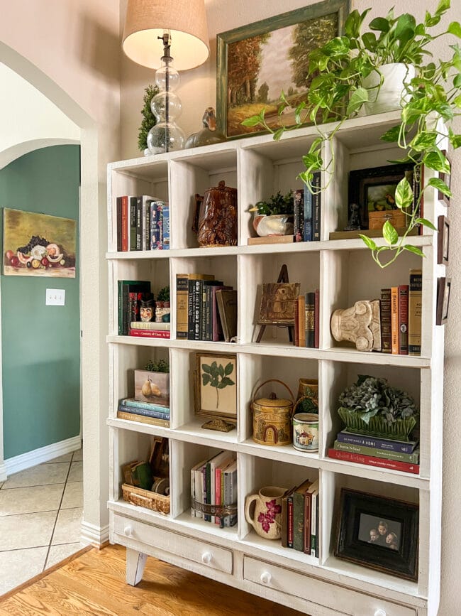 white cubby shelf with books, antiques with a plant on top next to a pastoral painting