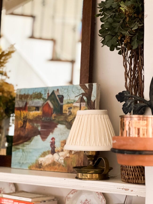 small lamp with white shade sitting in front of a brightly colored landscape photo and mirror