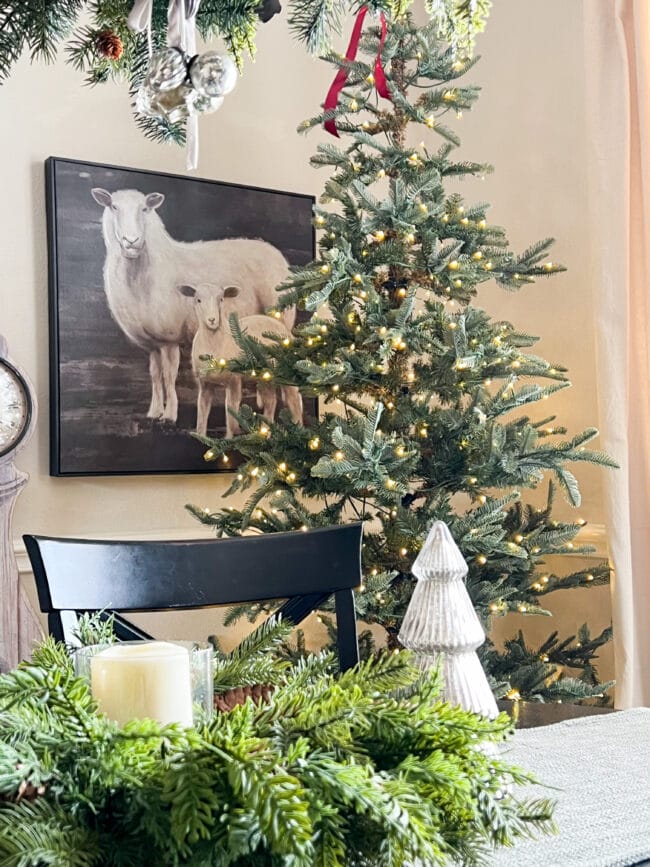 Christmas tree in the corner of room behind a table with a print of two lambs