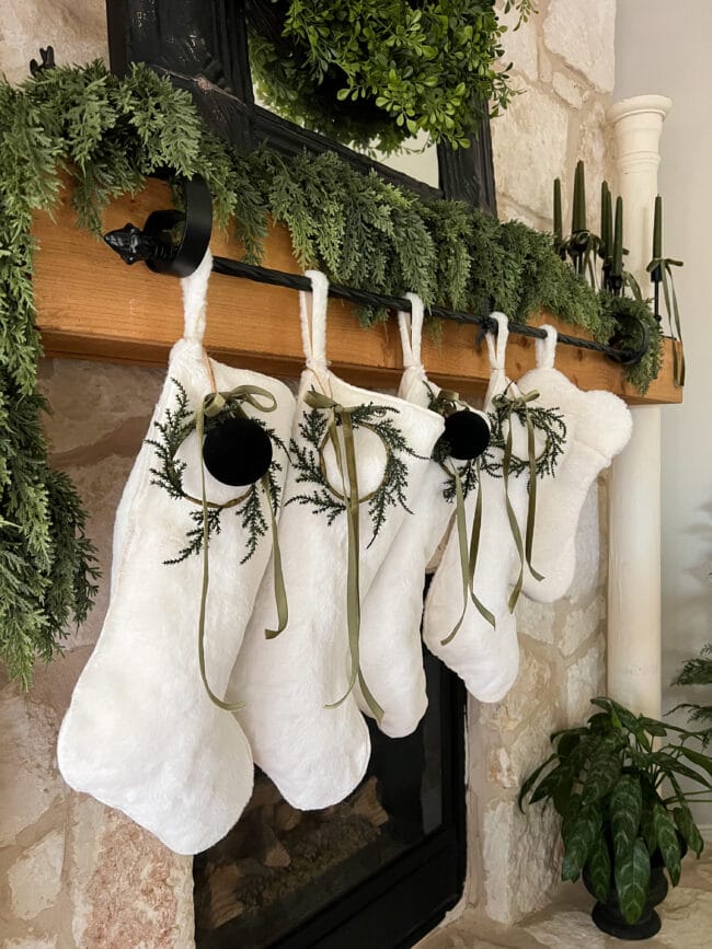 A row of white fur stockings hanging on a mantel. The stockings are decorated with black velvet balls, green ribbon and there's garland on top of the mantel