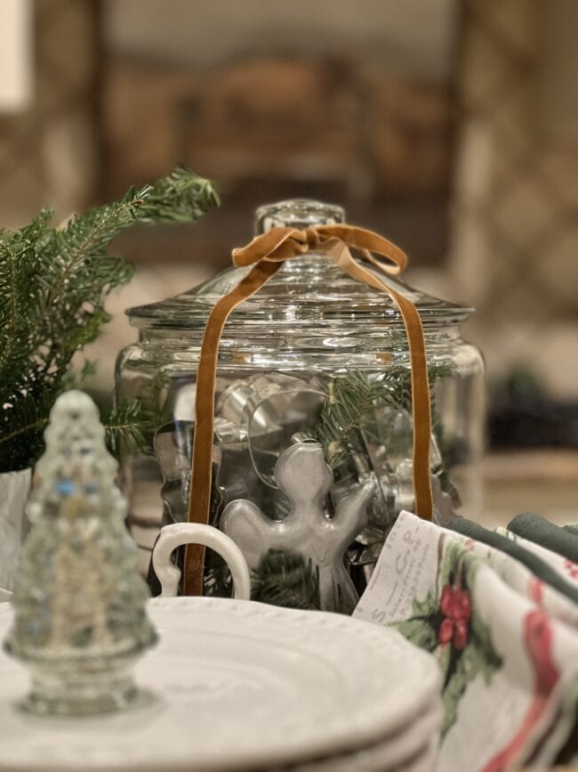 close up of glass jar with vintage Christmas cookie cutters, gold velvet bow on top and a small glass tree in the foreground.