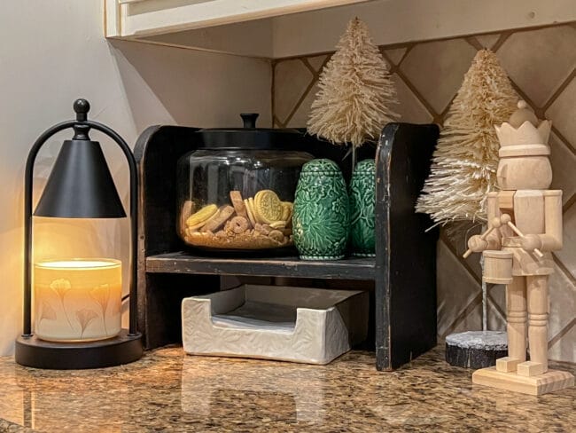 Black stool sitting in corner on kitchen counters wtih a candle warmer, cookie canister, green S&P shakers and white bottle brush trees 