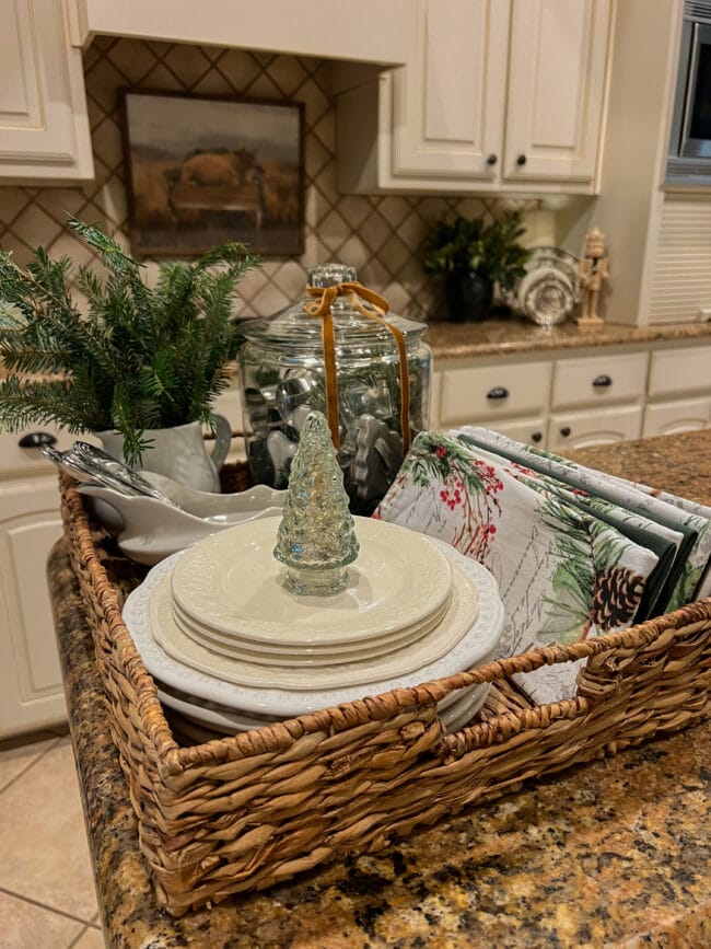 close up view of a basket tray with white plate, Christmas cloth napkins, a glass tree, pitcher with greenery and a glass canister with vintage cookie cutters inside