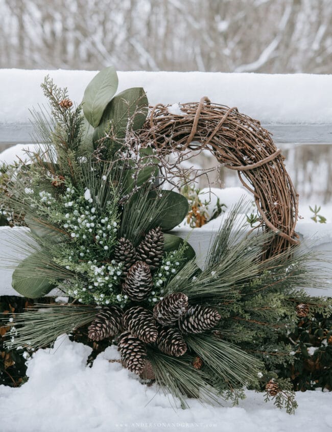 grapevine wreath surrounded by snow and has pinecones and winter stems on the wreath too