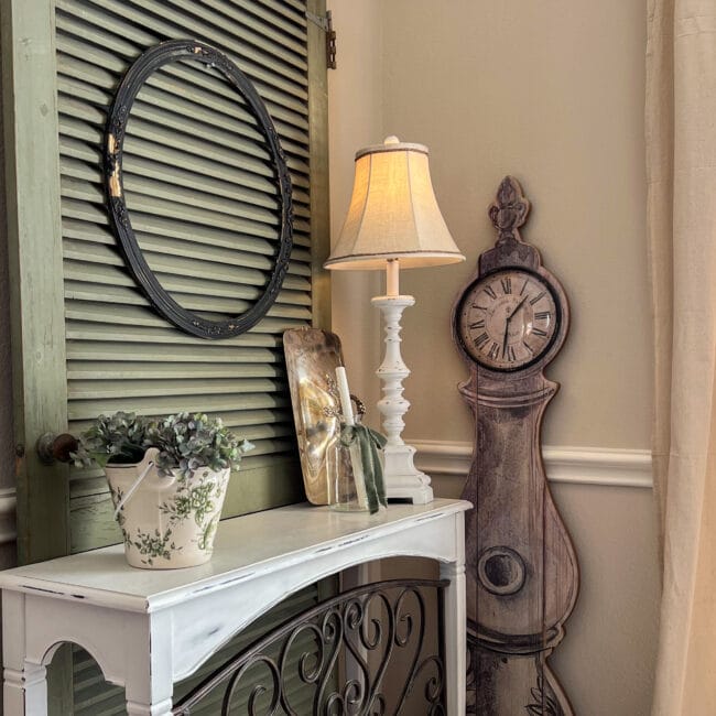 green screen door behind a white and iron table with a lamp, and candles. Floor clock leaning on wall to the right