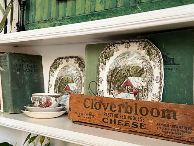 An old cloverbloom cheese box with books stacked inside and a vintage winter plate sitting in front 