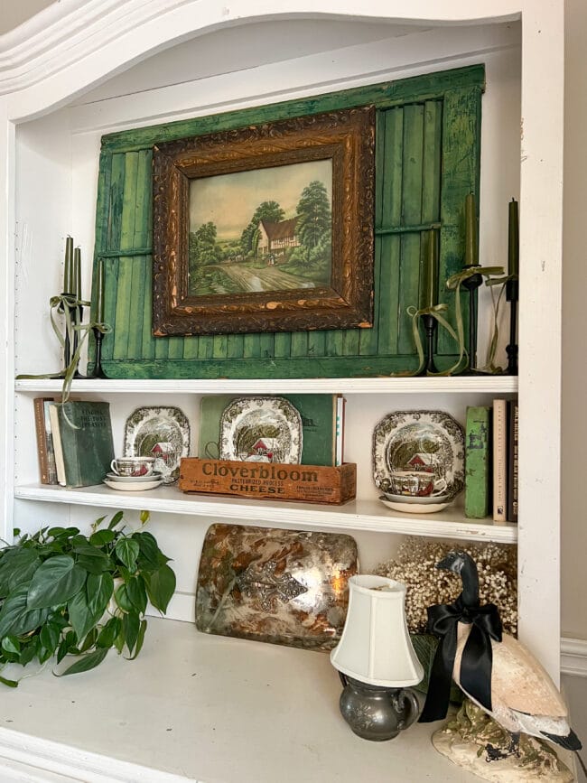 white hutch shelves with a green shutter, vintage print, candles, plates, books, and greenery
