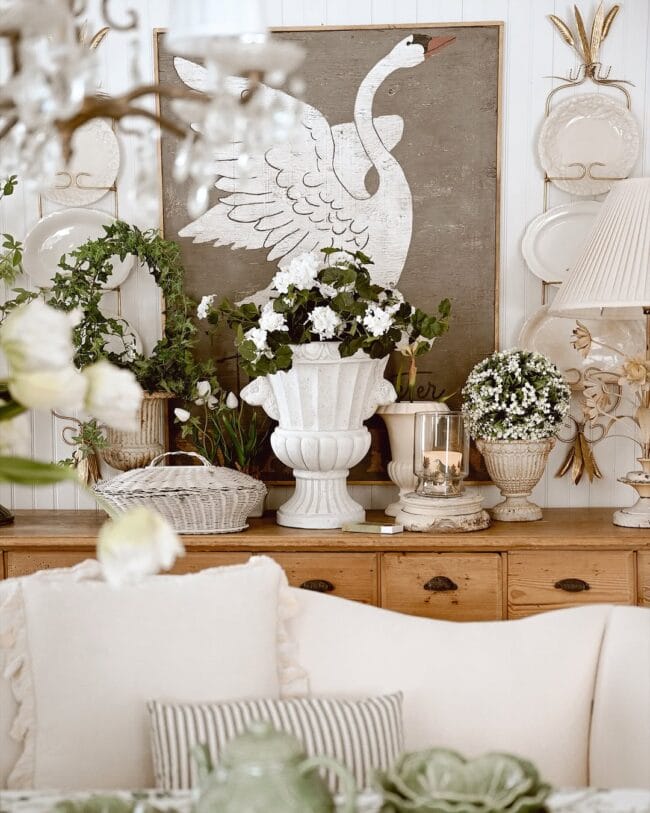 Spring Vignette with warm brown chest and on top are plants, white urns, plates on the wall and a large swan painting in background