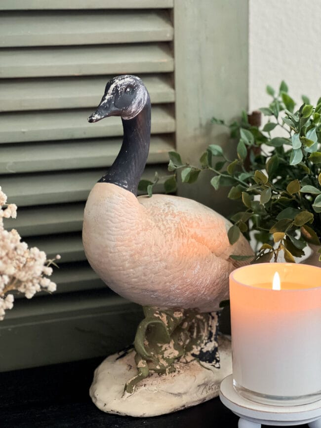 ceramic goose with a lit candle and plant beside it sitting in front of a vintage green wooden screen door