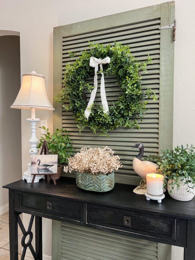 black console table with green door in back and a wreath on top of it. Geese, a lamp, greenery plants, and white Babys breath in the center