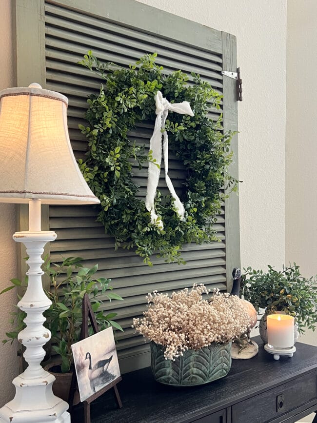 black console table with a vintage green screen door behind it. There's a white lamp, plants, a hanging wreath and a photo and a ceramic goose on the sides.