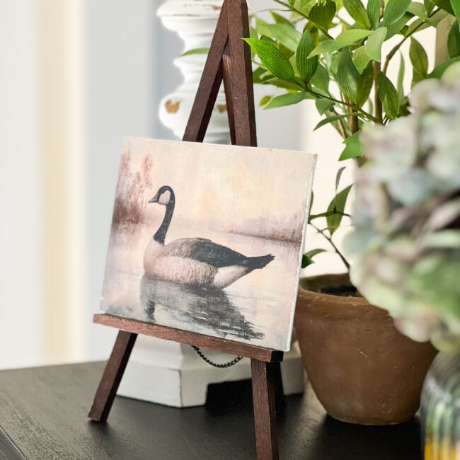 small painting of a goose on the water sitting on a tabletop easel with a plant in the background.