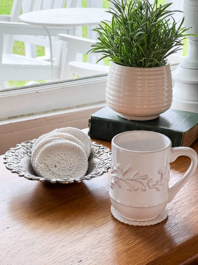 white coffee cup sitting on a wooden table with a white doily used as a coaster and several doilies in a silver bowl sitting next to it