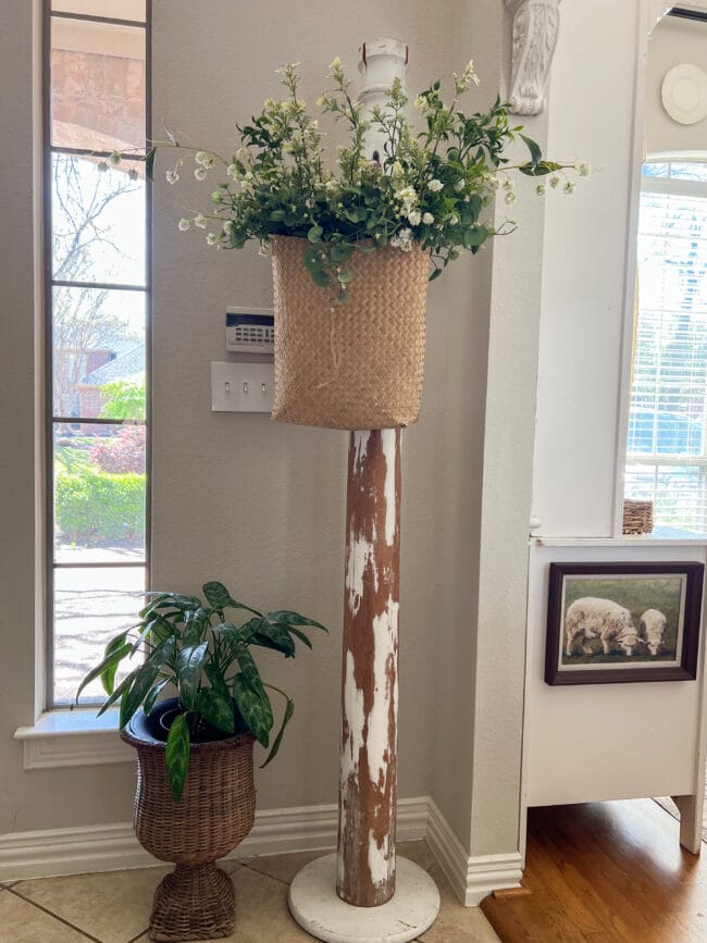 antique column made into a coat rack.  Hanging on it is a basket with faux spring stems and sitting beside it is a vintage wicker urn with plant