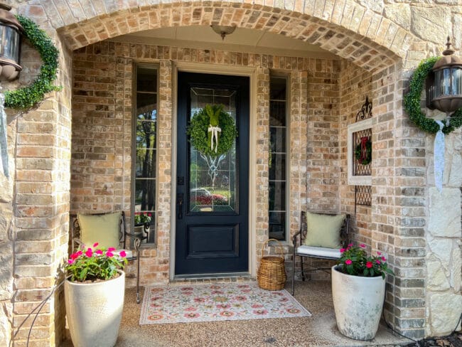 brick porch with two pots, chairs, a basket, rug and a wreath on the door with half of a rabbit 
