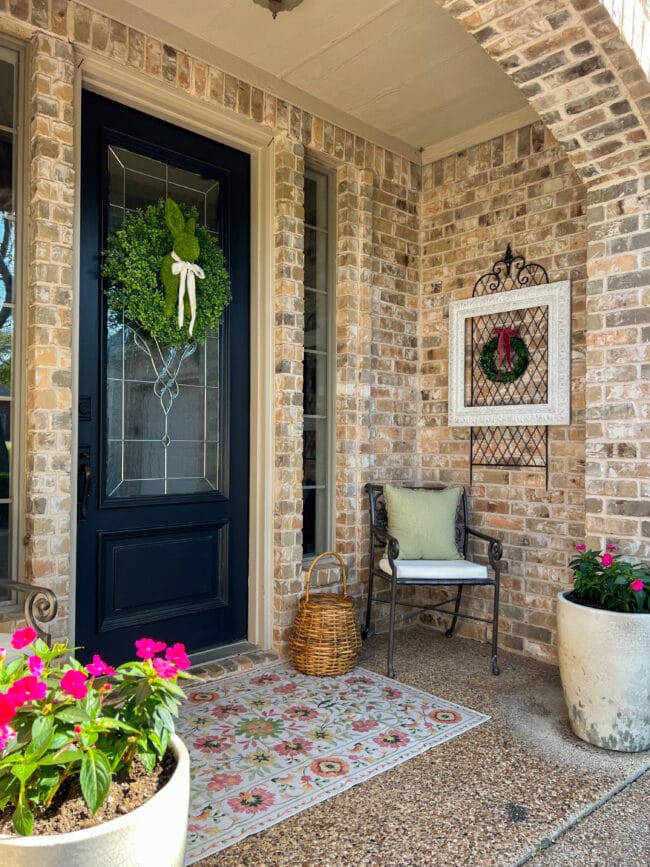 brick house with small porch. Boxwood wreath with rabbit in center, iron chair with green pillow, pink florals in pots, white frame hanging on brick and a colorful floral rug
