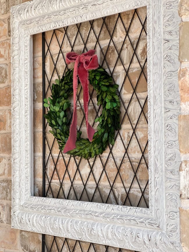 white frame with a small boxwood wreath and pink bow hanging inside on a brick wall