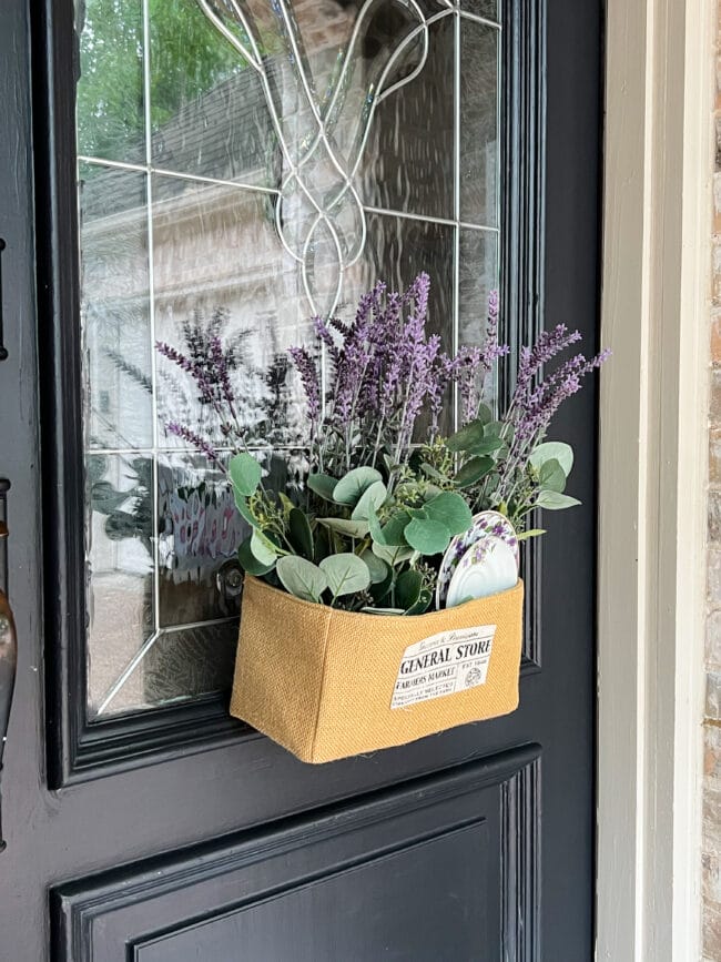close up image of a black front door with a burlap basket and purple flowers hanging on the glass 