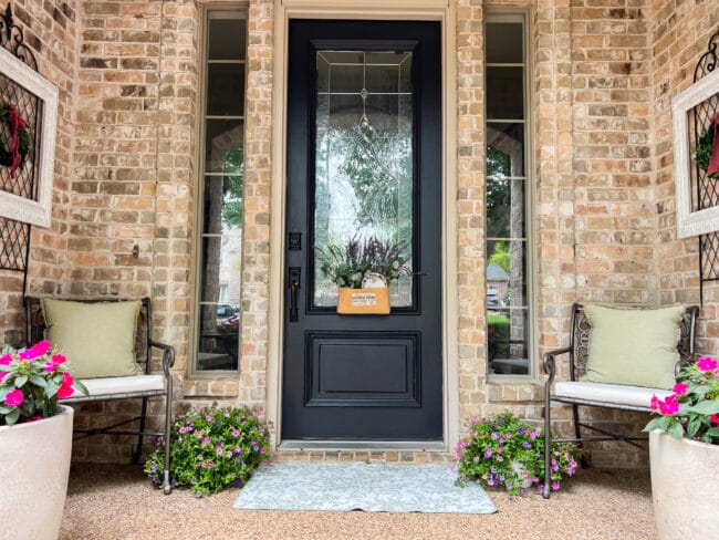 brick porch with black door, plants and two chairs with green pillows and white cushions