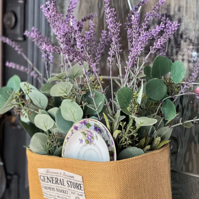 close up of a burlap basket with two vintage plates and faux purple and green flowers inside