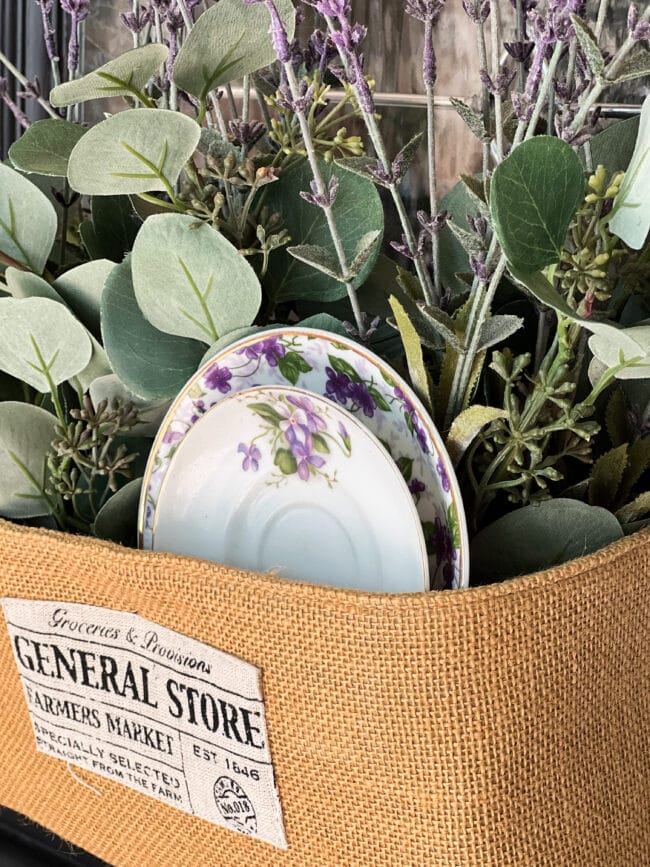 close up image of a burlap basket with greenery and two vintage plates with purple flowers