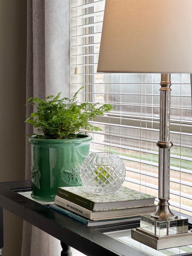 green planter with fern, lamp and candle on books sitting in front of a window with blinds