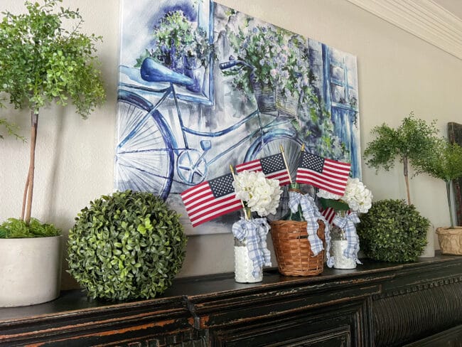 mantel with blue bike print, greenery, topiaries and 3 vases with flags and white flowers