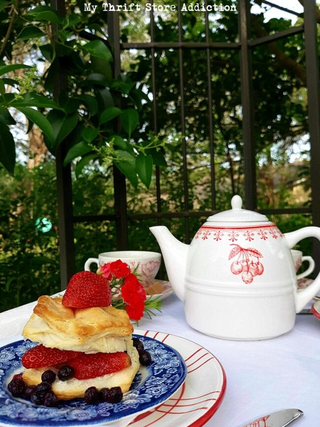 outdoor table set for two in a garden with patriot plates a teapot and a strawberry shortcake dessert.