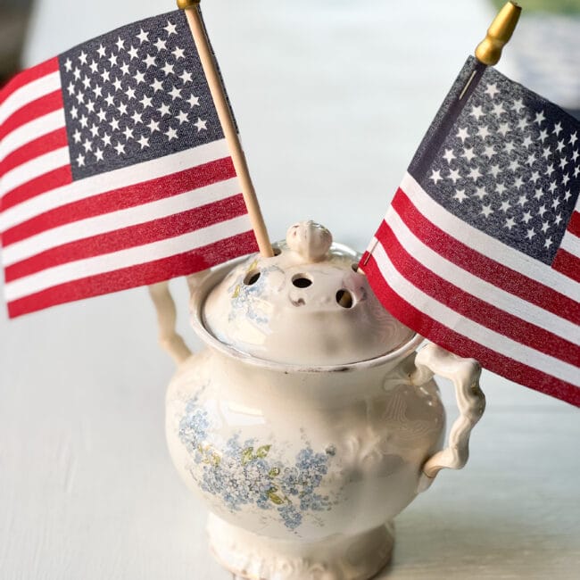 square photo of a vintage creamer with holes in the lid and 2 US flags stuck inside
