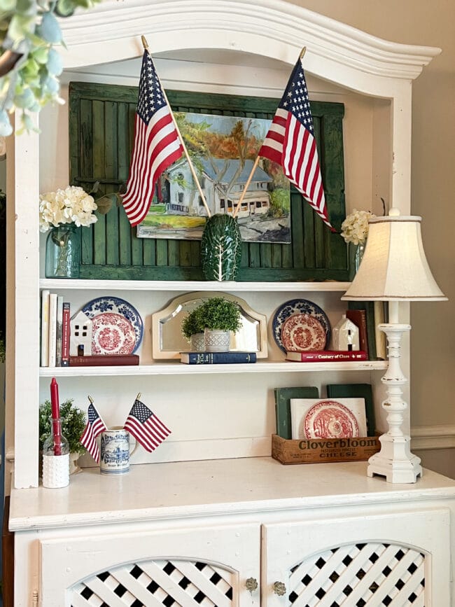 white hutch with green shutter and a painting of a farmhouse. Has flag, blue and white plates, books and a lamp on it