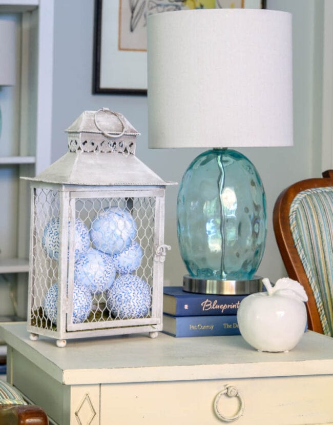 blue and white balls inside a white lantern sitting on a table with a blue lamp and a white ceramic apple