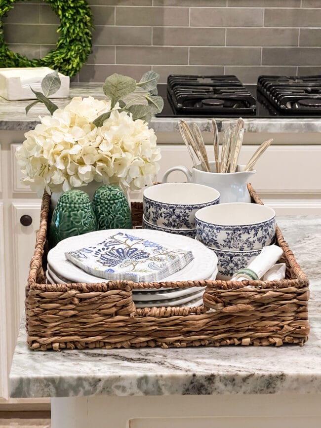 woven basket tray full of blue and white dishes, green s/p shakers, blue and green cocktail napkins and silverware and flowers in white pots