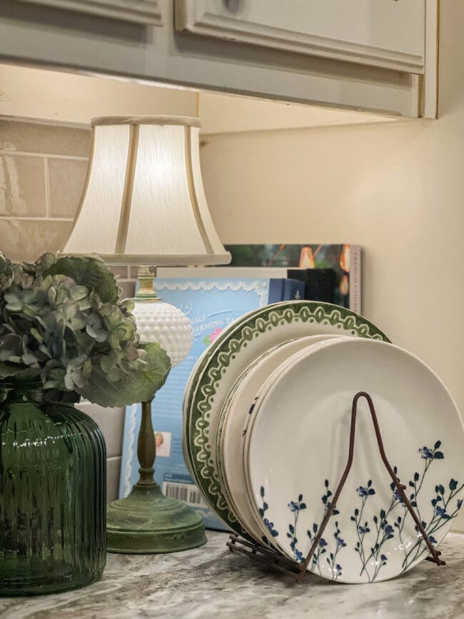 stack of plates in a corner with a vintage lamp, cookbooks and a green vase of faux hydrangeas