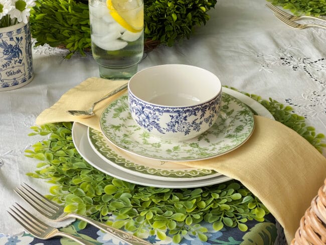 boxwood placemat with layered green and white dishes, a chinoiserie bowl and a yellow cloth napkin
