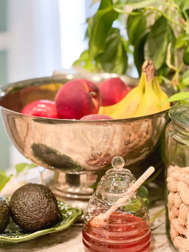 large silver plated bowl of fruit with a honey jar and avocados