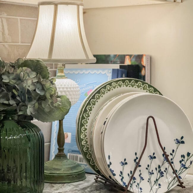plates, lamp, green vase with faux hydrangeas and cookbooks in the background