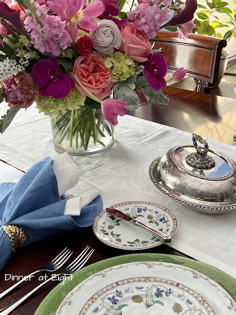 table setting with floral dishes, blue napkins and a pink floral arrangement