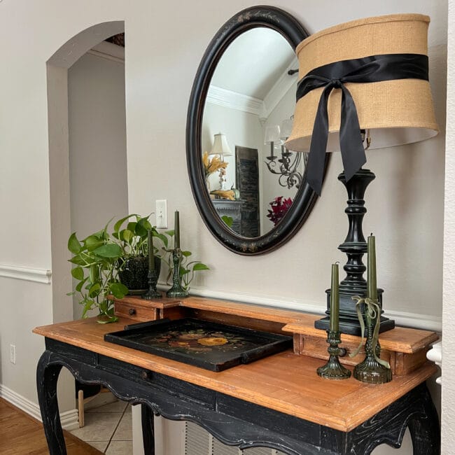 angled view of a two tone desk with an oval mirror, lamp with a black ribbon, plant, candles and a tole tray sitting on top.
