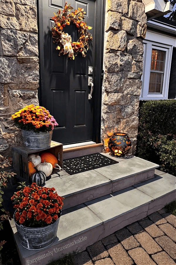 front porch with stone wall, wreath on black door, colorful mums in metal buckets, and a lit up lantern on the right side 
