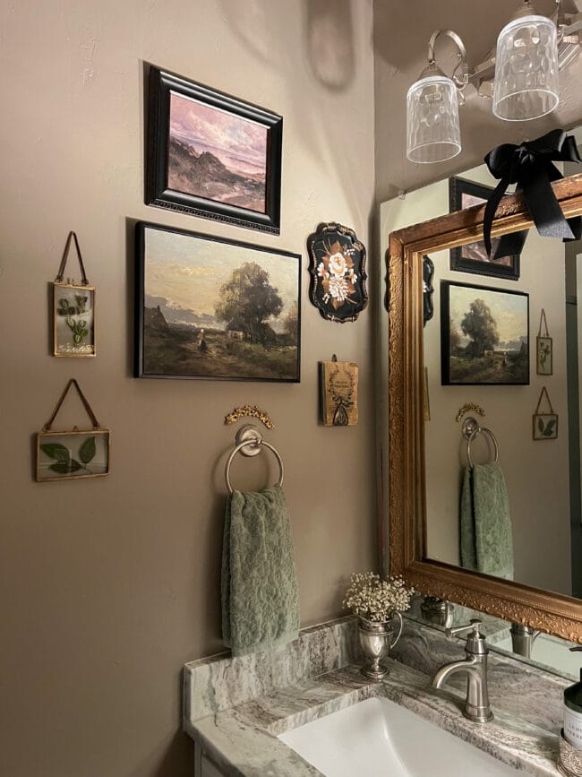 bathroom wall collage of small frames with leaves inside, hand towel, book on silver cake server and a small hand painted tray