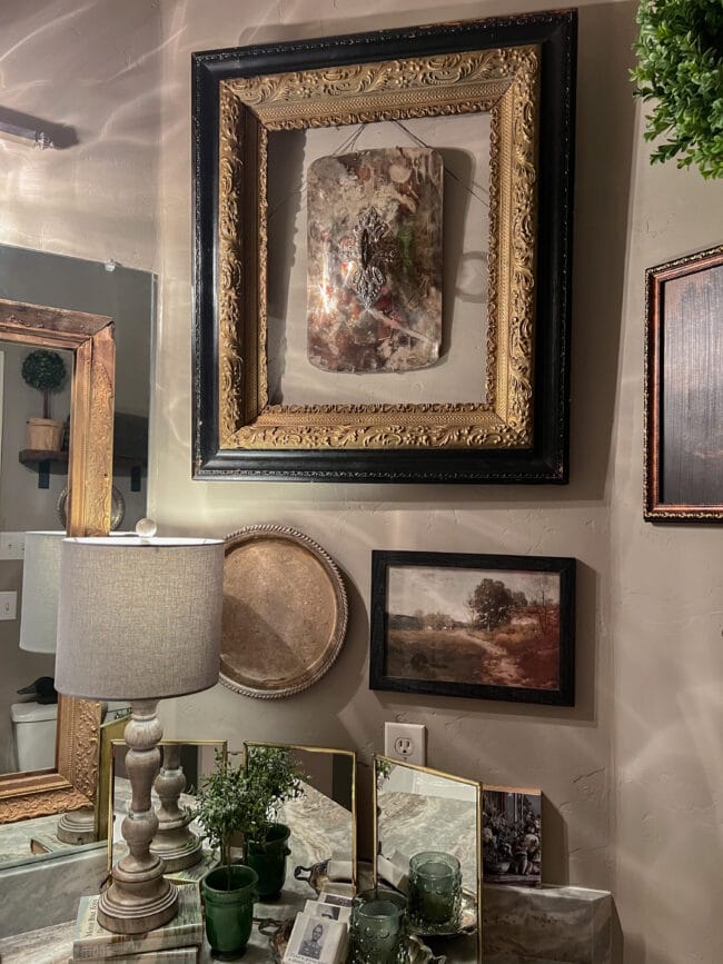 wall collage with paintings and a frame with a silver casserole lid hanging inside.