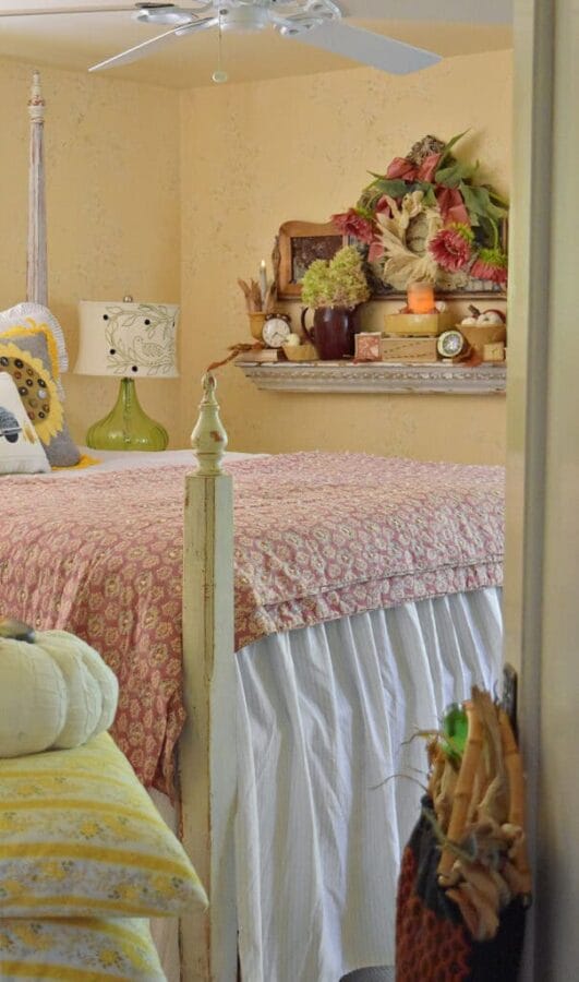 yellow walls in a bedroom with a poster bed and pink quilt. It has a lamp, and fall decor on the wall