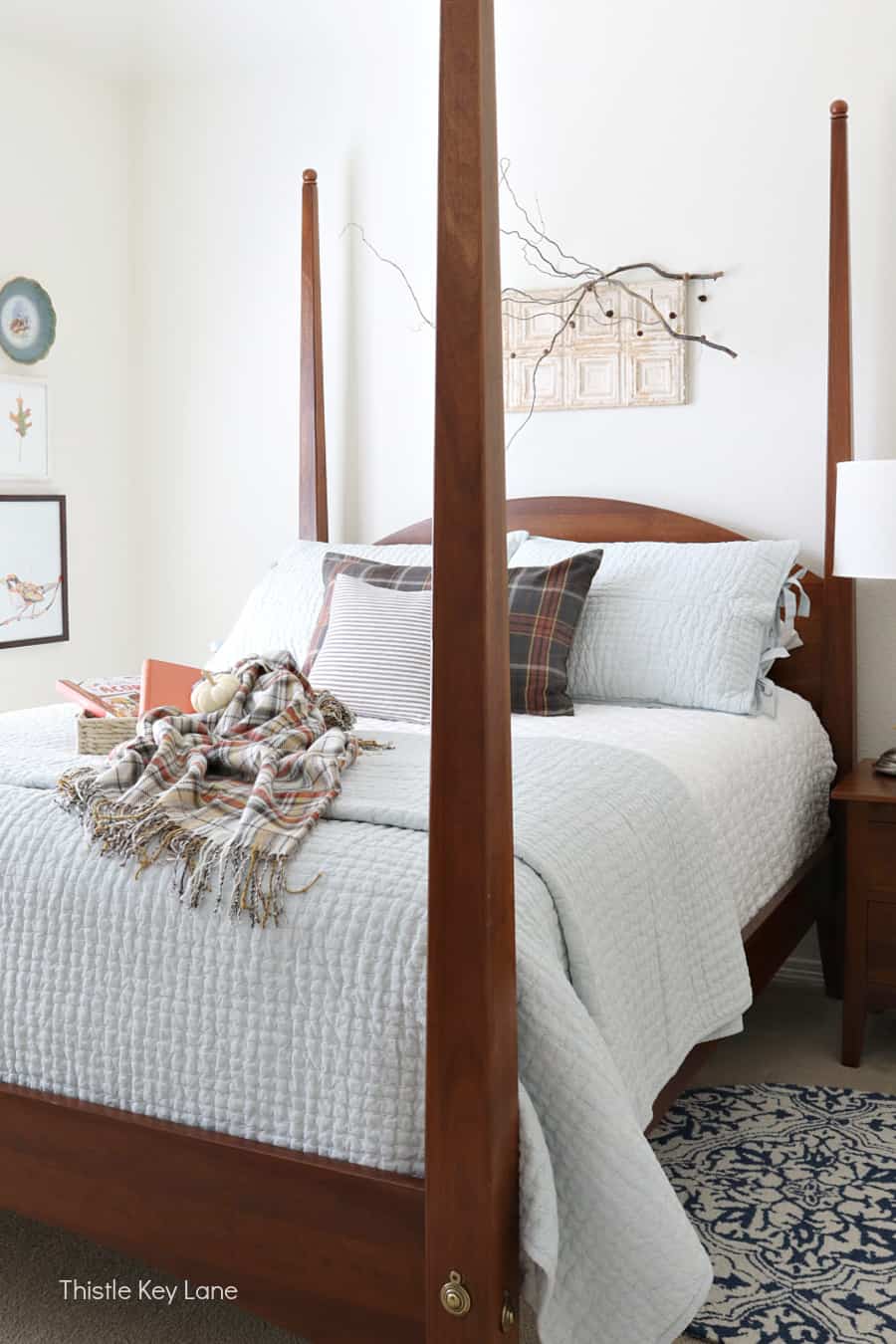 wooden 4 poster bed with white bedding, a plaid throw, tray with orange books and pumpkins on top of bed and a limb for artwork behind it.