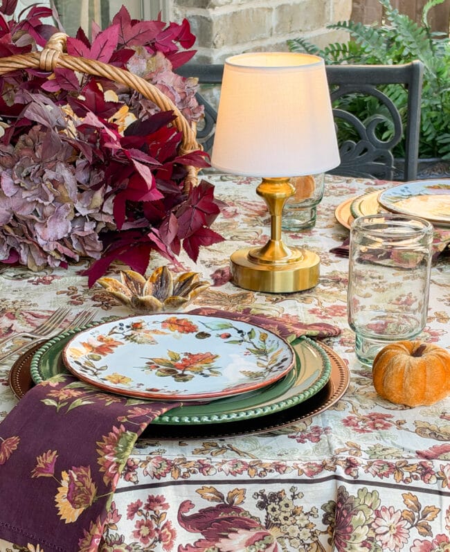 fall table with a lamp, autumn leafy basket and layered patterns with dishes in greens, burgundy and golds.