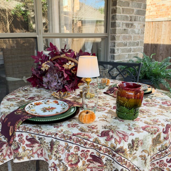 outdoor table setting with an autumn tablecloth, layered fall dishes, lamp and a basket with fall stems on the side