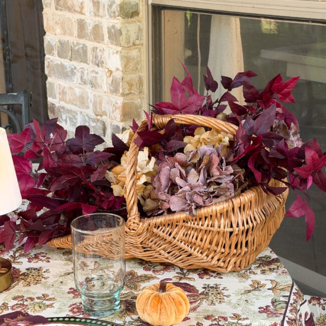 close up of a vintage basket with a handle and dark burgundy faux leafy stems and small gold and purple faux hydrangeas